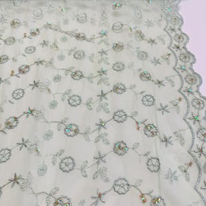 Peut inclure: Un rouleau de tissu tulle vert clair d&eacute;licat avec une broderie florale complexe. Le motif pr&eacute;sente des fils argent&eacute;s soulignant des fleurs et des feuilles, agr&eacute;ment&eacute; de paillettes iris&eacute;es. Le bord du tissu a une bordure festonn&eacute;e.