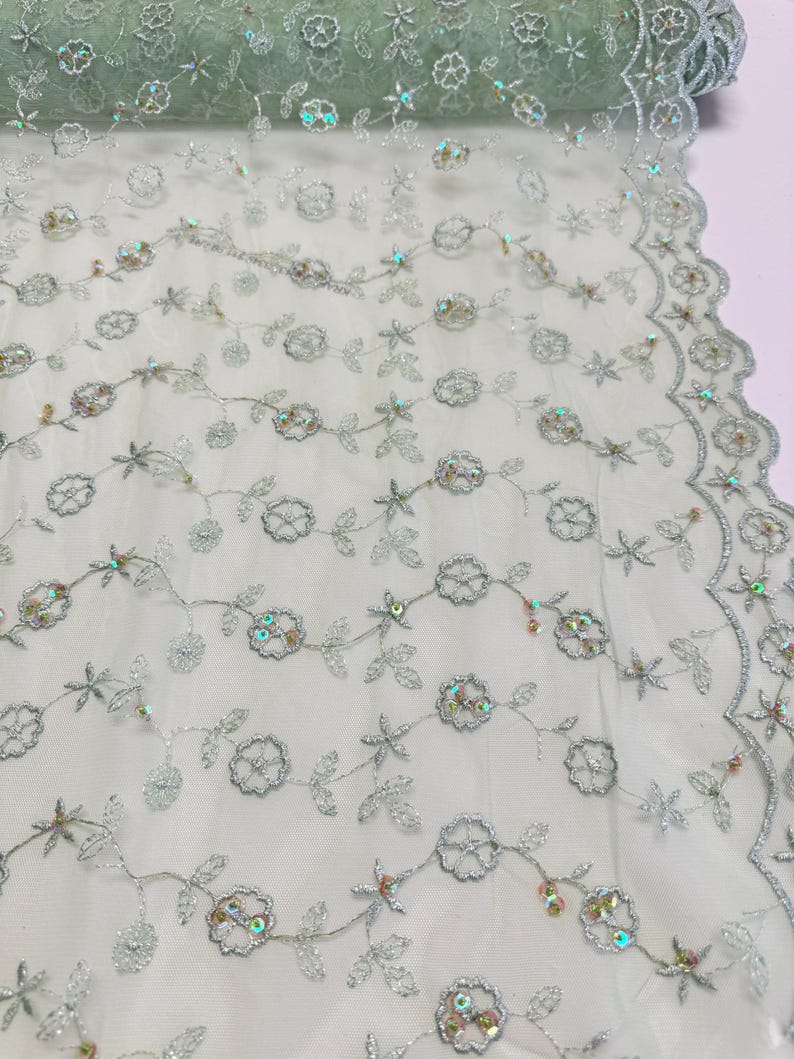 Peut inclure: Un rouleau de tissu tulle vert clair d&eacute;licat avec une broderie florale complexe. Le motif pr&eacute;sente des fils argent&eacute;s soulignant des fleurs et des feuilles, agr&eacute;ment&eacute; de paillettes iris&eacute;es. Le bord du tissu a une bordure festonn&eacute;e.