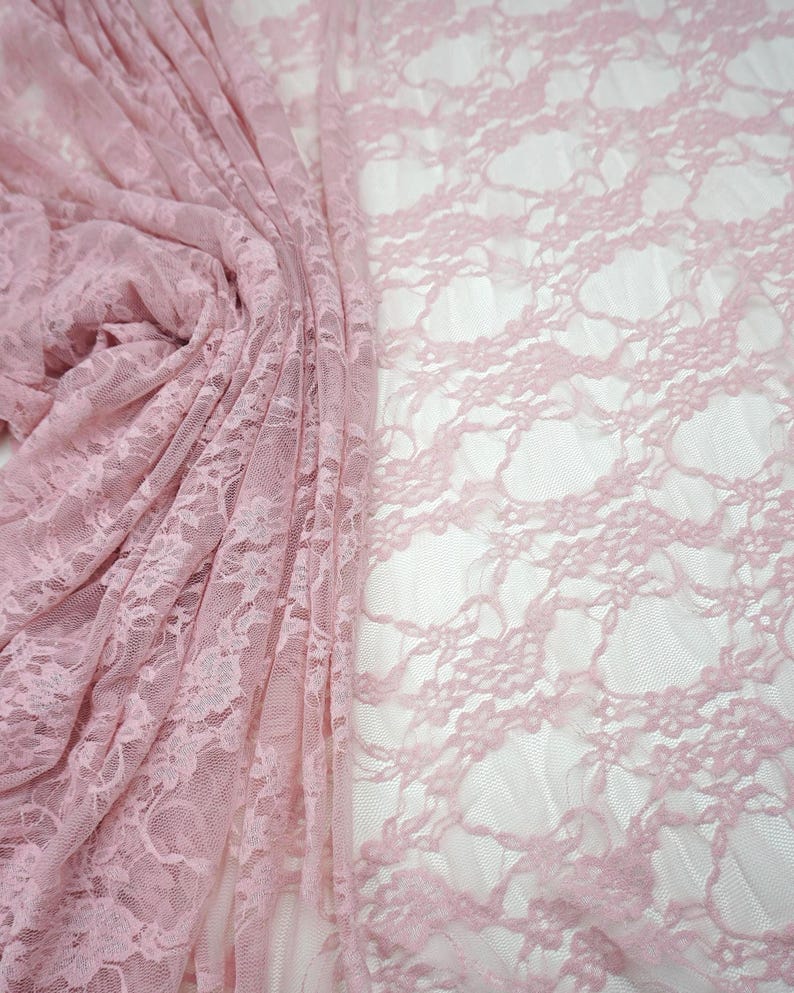 May include: A close-up of delicate, light pink lace fabric. The fabric is draped and folded, revealing intricate floral patterns. The lace has a soft, airy appearance, perfect for various craft projects or apparel.
