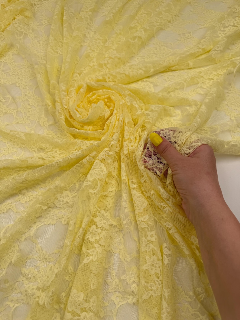 K&ouml;nnte beinhalten: Nahaufnahme eines Wirbels aus gelbem Spitzenstoff. Das zarte Material ist mit floralen Mustern versehen. Der Stoff wird von einer Hand mit gelbem Nagellack gehalten.