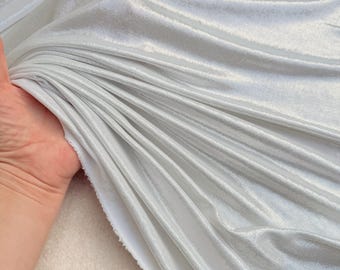 May include: A piece of shimmering silver fabric with a metallic finish and a soft, velvet-like texture. The material is draped in deep, flowing folds that catch the light, highlighting the reflective surface and the smooth, consistent sheen.