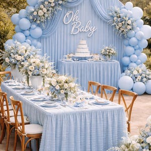 Op de afbeelding: Een babyborrel ingericht in lichtblauw. De sc&egrave;ne toont een tafelsetting met bloemstukken, een taart en een achtergrond met ballonnen en de tekst "Oh Baby". Houten stoelen en een lange tafel zijn gedekt voor de gasten.