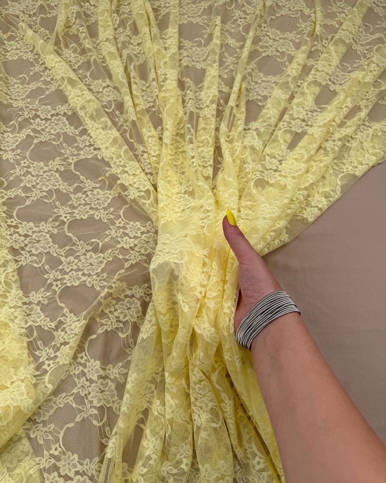 May include: Pale yellow lace fabric with a floral pattern. The delicate material is draped, revealing the intricate design. The fabric is held by a hand wearing a silver bracelet. The background is a neutral beige.