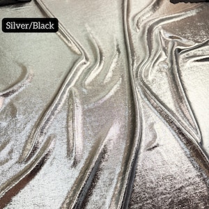 May include: A piece of fabric with a metallic silver finish on a black base, featuring a draped texture. A black label in the upper left corner reads Silver/Black, and text at the bottom states Multiple yards will come in one piece.