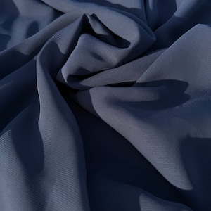 Può includere: Primo piano di un tessuto blu scuro con una superficie liscia e leggermente strutturata. Il tessuto è drappeggiato e piegato, creando linee morbide e fluide.