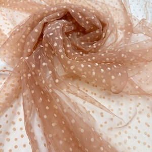 May include: Close-up of a sheer, peach-coloured fabric with a polka dot pattern. The fabric is gathered and folded, creating a layered effect. The white polka dots are evenly spaced across the material.