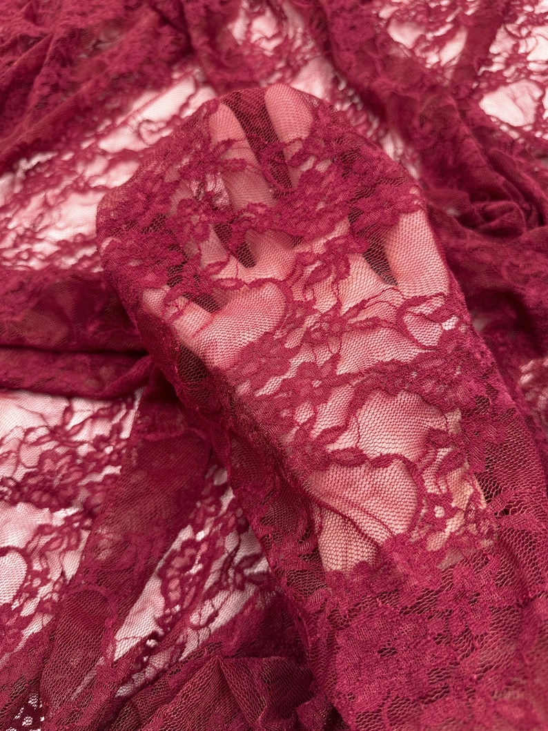 K&ouml;nnte beinhalten: Nahaufnahme von bordeauxfarbenem Spitzenstoff. Der zarte Stoff weist ein kompliziertes Blumenmuster auf. Das transparente Material ist drapiert und zeigt die Textur und das Design der Spitze. Die Farbe ist ein tiefer, satter Rotton.