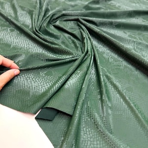 Puede incluir: Tela verde oscuro con un estampado de piel de serpiente. El material está drapeado y doblado, mostrando la textura y el brillo de la tela. Una mano toca la tela, resaltando su textura.