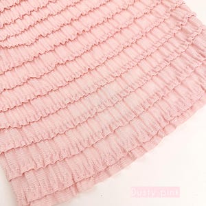 Op de afbeelding: Stof in stoffig roze met gelaagde ruches. De stof heeft een getextureerde uitstraling en is lichtroze van kleur. De tekst "Dusty pink" en "Multiple yards will come in one piece" zijn zichtbaar.