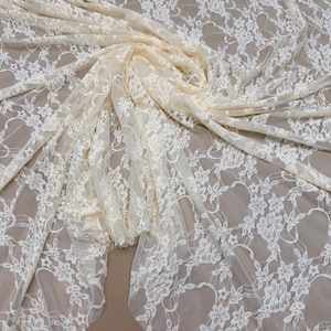 May include: Cream-colored lace fabric with a floral pattern. The fabric is draped and folded, showcasing its texture and design. The text "Cream" is at the top left, and "58/60 inches wide" is at the bottom.