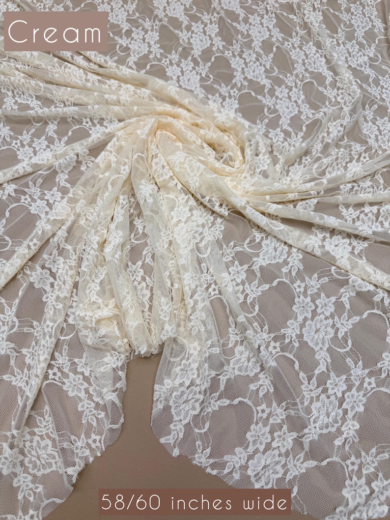 May include: Cream-colored lace fabric with a floral pattern. The fabric is draped and folded, showcasing its texture and design. The text "Cream" is at the top left, and "58/60 inches wide" is at the bottom.