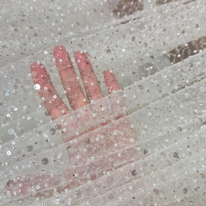 May include: A sheer white tulle fabric embellished with a dense arrangement of small white pearls, clear sequins, and metallic silver beads. The lightweight mesh material features a repeating pattern of these decorative elements across the surface.