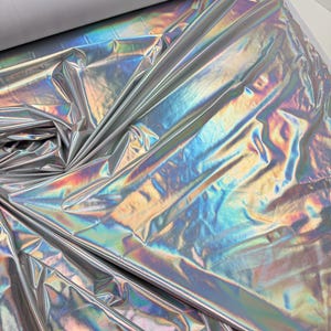 Puede incluir: Tela plateada iridiscente con brillo metálico. La tela refleja un arcoíris de colores, creando un efecto brillante. Un rollo de tela blanca es visible en el fondo. La tela se utiliza probablemente para ropa, disfraces o fines decorativos.