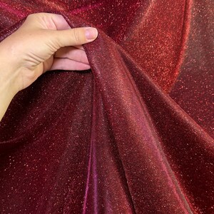 May include: Close-up of a burgundy fabric with a sparkly, iridescent finish. The fabric is soft and flowing, with a subtle sheen.