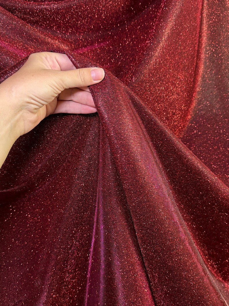 May include: Close-up of a burgundy fabric with a sparkly, iridescent finish. The fabric is soft and flowing, with a subtle sheen.