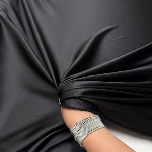 May include: A large piece of black satin fabric, draped and folded, creating deep shadows and highlights. The fabric has a smooth, lustrous surface. The image shows the fabric's texture and sheen, ideal for apparel or home decor projects.