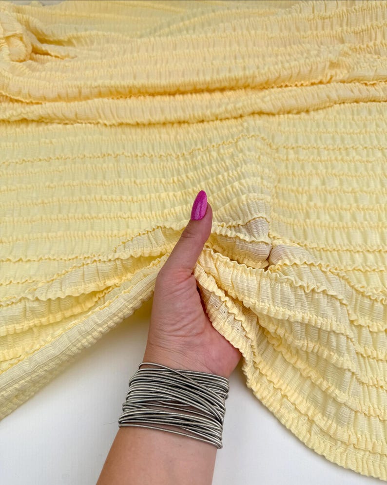 Op de afbeelding: Een lichtgele, getextureerde stof met horizontale shirring wordt vastgehouden door een hand met een zilveren armband. De stof heeft een gekreukeld uiterlijk. De hand heeft een roze gelakte nagel.