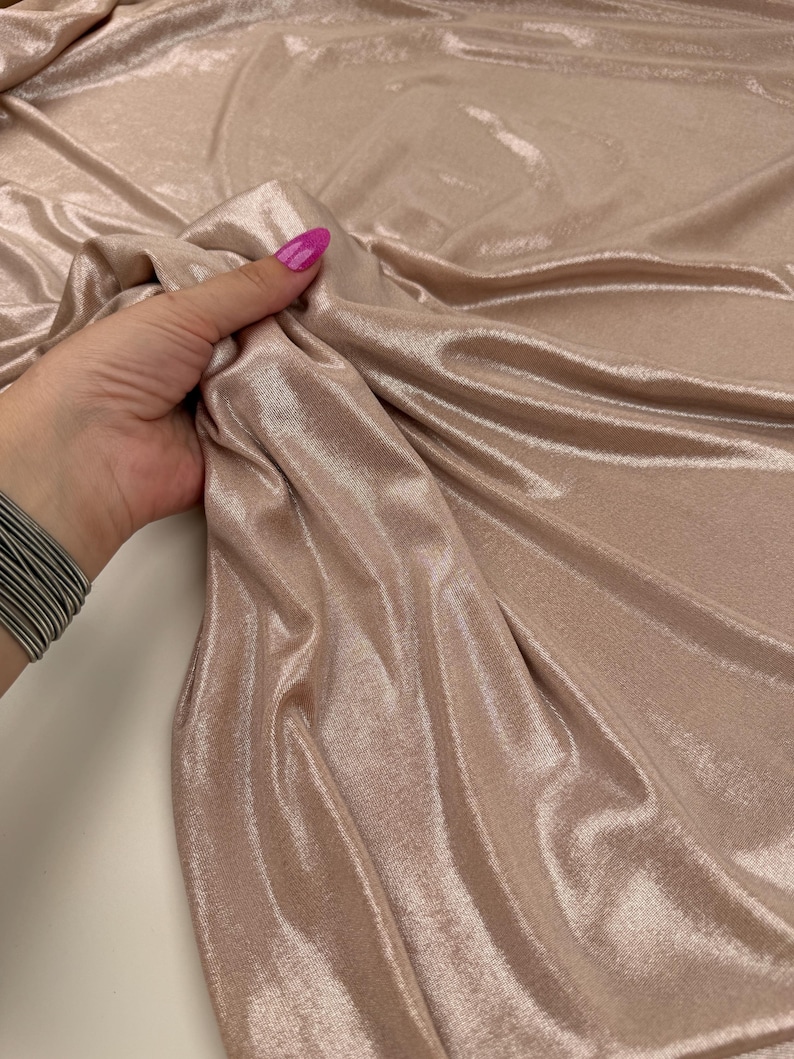 May include: A shimmering, light champagne-coloured fabric with a metallic finish and a smooth, reflective surface. The material is draped in soft, flowing folds, highlighting its lustrous texture and consistent, light-reflecting quality throughout.