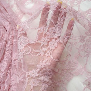 May include: Close-up of delicate pink lace fabric with floral patterns. The sheer material is held up, revealing the intricate design and texture. The lace is a soft, muted pink, ideal for apparel or craft projects.