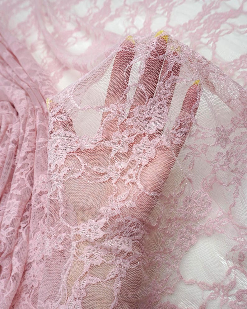 May include: Close-up of delicate pink lace fabric with floral patterns. The sheer material is held up, revealing the intricate design and texture. The lace is a soft, muted pink, ideal for apparel or craft projects.