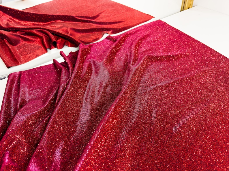 May include: Red sparkly fabric with a textured, glittery surface. The fabric is folded and draped, showing the shimmery texture.