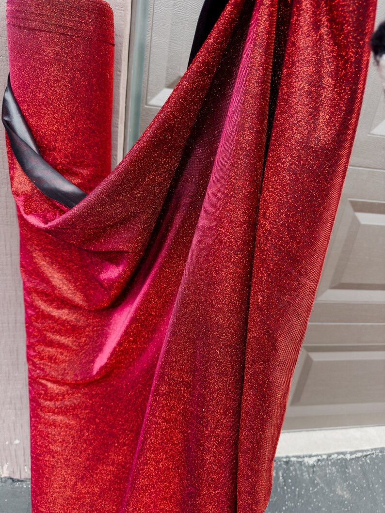 May include: Red sparkly fabric with a slight ombre effect. The fabric is rolled up and draped over a white surface.