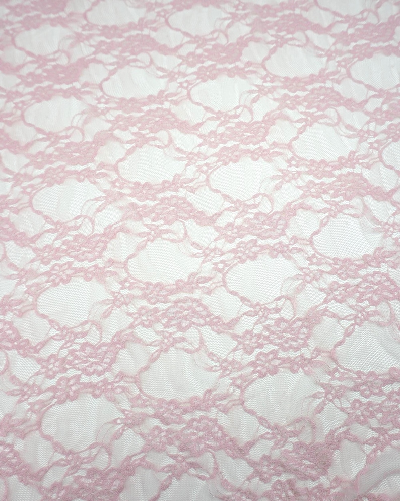 May include: Close-up of a light pink lace fabric. The delicate lace features a floral pattern with a repeating design. The fabric is semi-transparent, with a soft, textured appearance. The background is white.