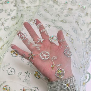 Peut inclure: Tissu vert clair et transparent avec broderies florales et paillettes iris&eacute;es. Le motif pr&eacute;sente des fleurs et des feuilles en fil d'argent. Le tissu est translucide, cr&eacute;ant un effet de superposition. Id&eacute;al pour les robes, les voiles ou les projets d&eacute;coratifs. Mesures en centim&egrave;tres.