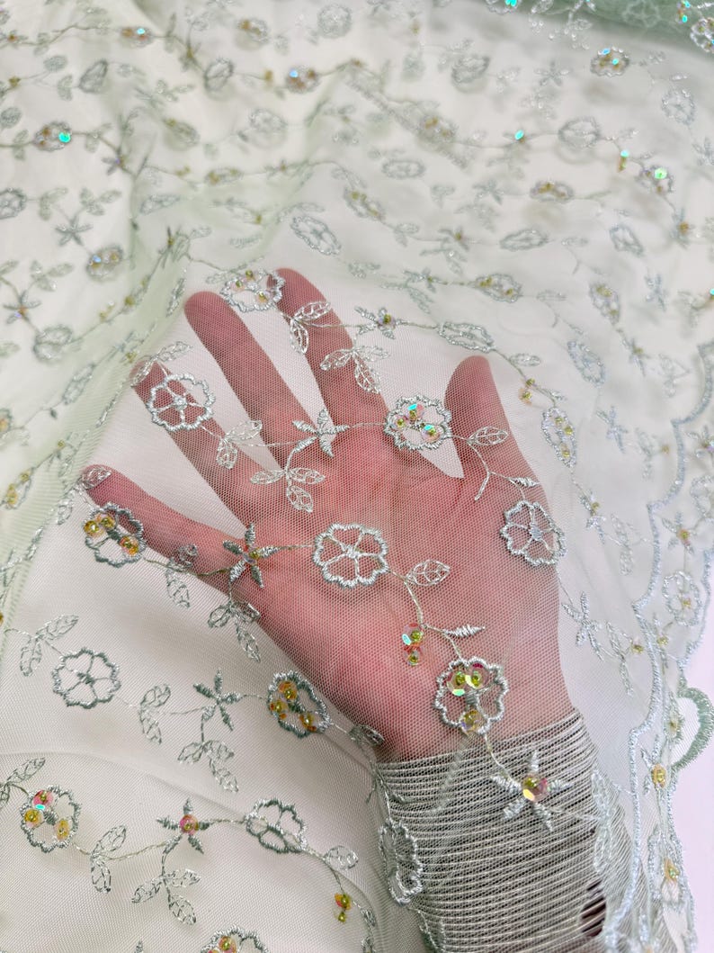 Peut inclure: Tissu vert clair et transparent avec broderies florales et paillettes iris&eacute;es. Le motif pr&eacute;sente des fleurs et des feuilles en fil d'argent. Le tissu est translucide, cr&eacute;ant un effet de superposition. Id&eacute;al pour les robes, les voiles ou les projets d&eacute;coratifs. Mesures en centim&egrave;tres.