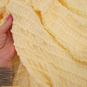Op de afbeelding: Close-up van een lichtgele stof met een getextureerd, gerimpeld ontwerp. De stof wordt vastgehouden door een hand met een roze gelakte nagel. Het woord "Yellow" staat in een gele doos linksonder.