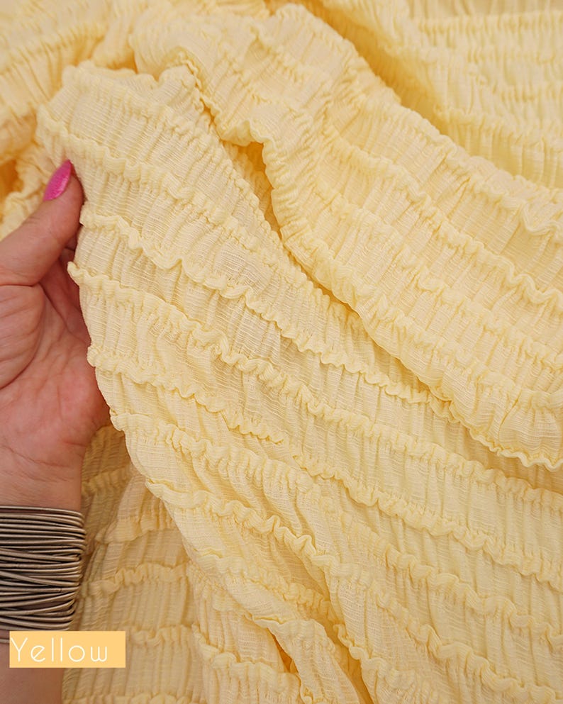 Op de afbeelding: Close-up van een lichtgele stof met een getextureerd, gerimpeld ontwerp. De stof wordt vastgehouden door een hand met een roze gelakte nagel. Het woord "Yellow" staat in een gele doos linksonder.