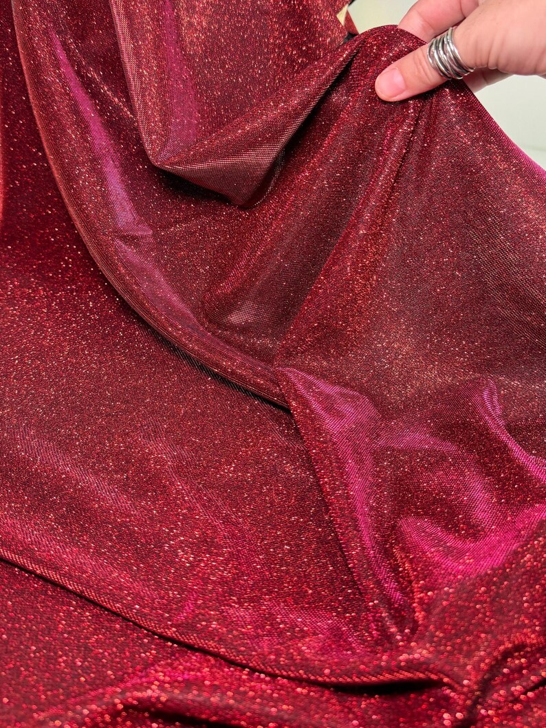 May include: Red sparkly fabric with a shimmery finish. The fabric has a fine, textured weave and a subtle sheen.