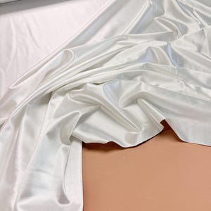 K&ouml;nnte beinhalten: Wei&szlig;er Satin-Stoff mit einer glatten, seidenartigen Textur. Der Stoff ist drapiert und gefaltet und zeigt seine luxuri&ouml;se Haptik.