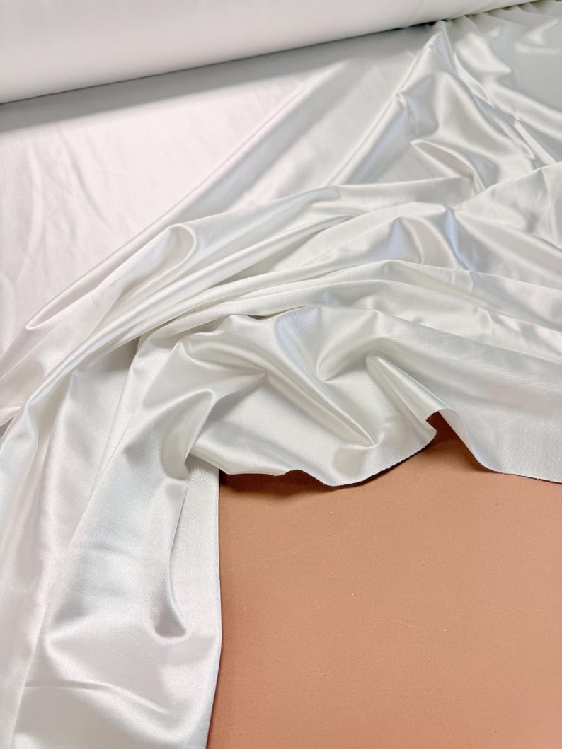 K&ouml;nnte beinhalten: Wei&szlig;er Satin-Stoff mit einer glatten, seidenartigen Textur. Der Stoff ist drapiert und gefaltet und zeigt seine luxuri&ouml;se Haptik.