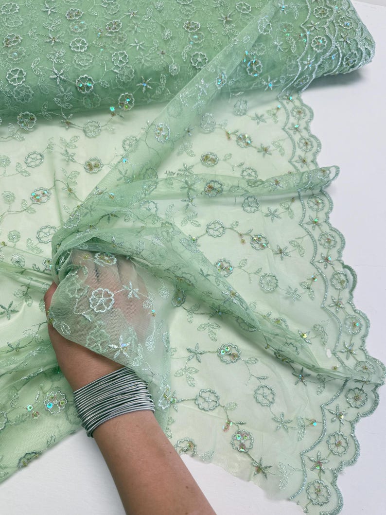 Peut inclure: Tissu en dentelle vert clair avec des fleurs brod&eacute;es et des paillettes. Le tissu d&eacute;licat est pli&eacute;, r&eacute;v&eacute;lant les d&eacute;tails complexes du motif. La dentelle a un bord festonn&eacute;. Id&eacute;al pour la couture.