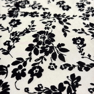 Op de afbeelding: Witte stof met een zwart bloemenpatroon. De bloemen zijn klein en delicaat, en het patroon wordt over de hele stof herhaald.