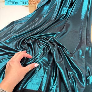 May include: A length of metallic fabric in a deep teal shade, labelled as Tiffany blue. The material features a highly reflective, shimmering surface with a smooth texture that drapes in soft folds, revealing a solid black backing underneath.