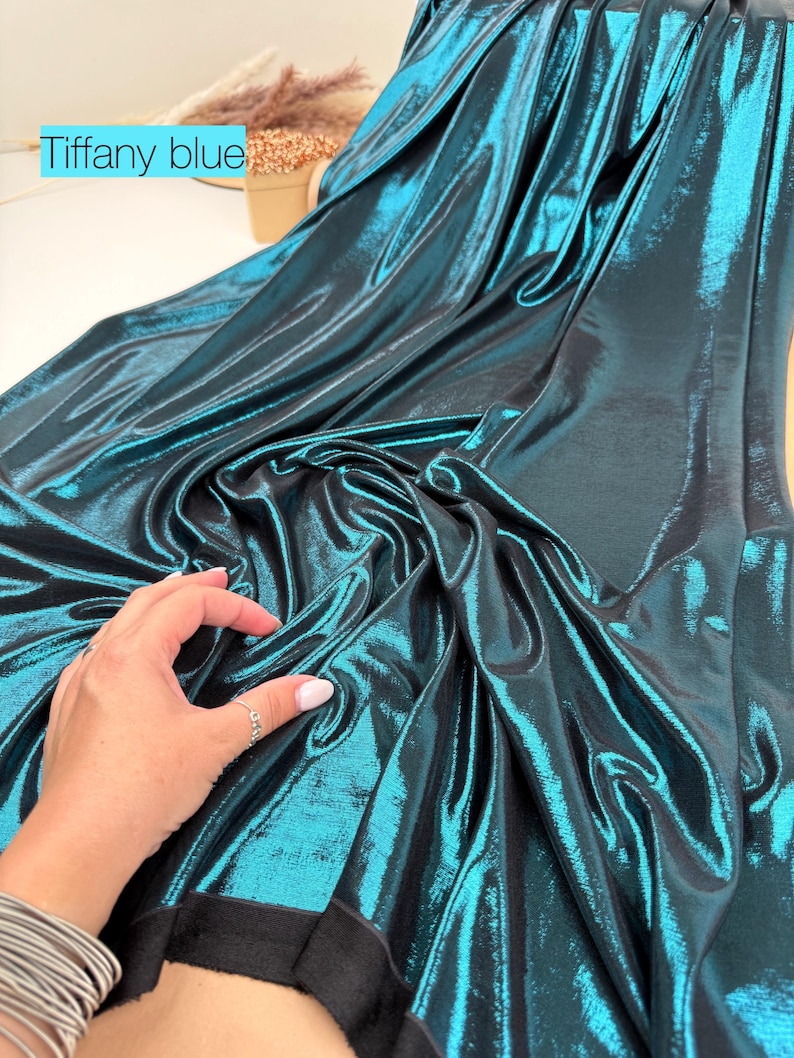 May include: A length of metallic fabric in a deep teal shade, labelled as Tiffany blue. The material features a highly reflective, shimmering surface with a smooth texture that drapes in soft folds, revealing a solid black backing underneath.