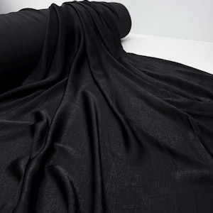 May include: A roll of black fabric with a textured surface. The fabric is draped, showing folds and shadows. The material appears to be a flowing, lightweight textile, suitable for apparel or decorative projects.