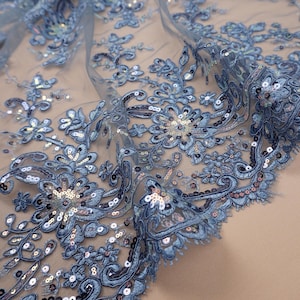 Könnte beinhalten: Nahaufnahme eines zarten blauen Spitzenstoffs mit floralen Stickereien und schimmernden Pailletten. Das aufwendige Design zeigt detaillierte Blumenmuster und wirbelnde Verzierungen, wodurch ein luxuriöses und elegantes Textil entsteht.