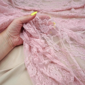 Może przedstawiać: Zbliżenie delikatnej różowej koronki z kwiatowymi wzorami. Przezroczysty materiał jest trzymany, ukazując skomplikowane detale i fakturę. Koronka jest w kolorze miękkiego różu, idealna do projektów odzieżowych lub rzemieślniczych.