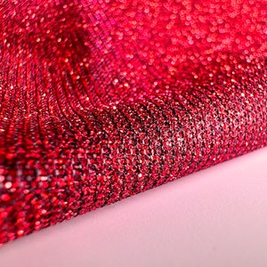 May include: Red glitter fabric with a textured, sparkly surface. The fabric is folded in half, showing the shiny, iridescent texture.