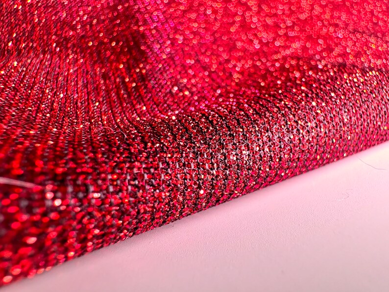 May include: Red glitter fabric with a textured, sparkly surface. The fabric is folded in half, showing the shiny, iridescent texture.