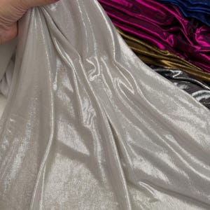 May include: A piece of silver metallic fabric with a high-shine, reflective finish. The material is draped to show its fluid texture and light-catching surface. Additional rolls of fabric in blue, magenta, gold, and dark grey are visible behind.