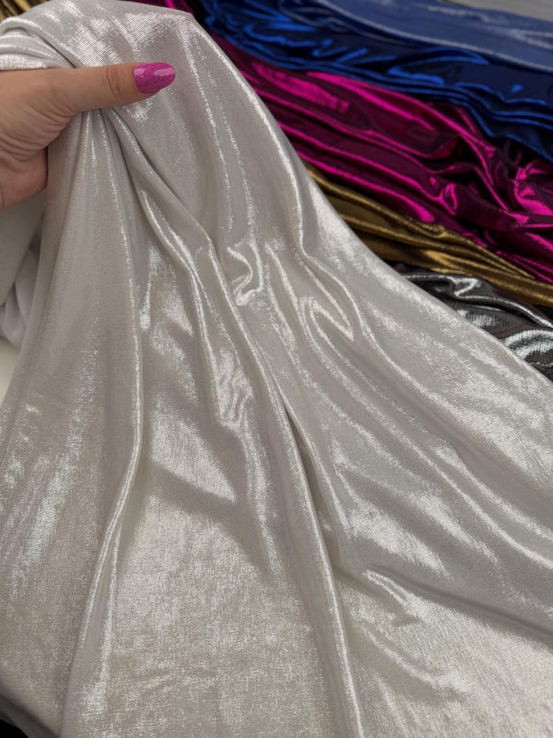 May include: A piece of silver metallic fabric with a high-shine, reflective finish. The material is draped to show its fluid texture and light-catching surface. Additional rolls of fabric in blue, magenta, gold, and dark grey are visible behind.
