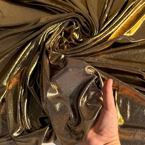 May include: A metallic gold fabric with a shimmering, reflective surface and a fine-grained texture. The material is draped in soft, flowing folds, creating deep shadows and bright highlights that emphasise its lustrous, liquid-like appearance.