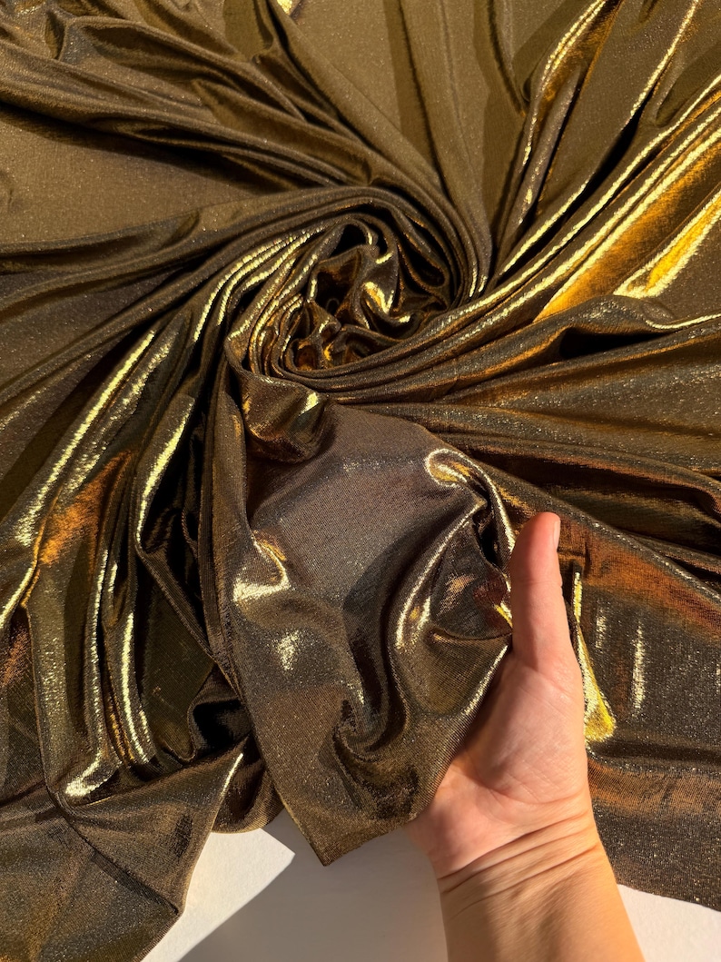 May include: A metallic gold fabric with a shimmering, reflective surface and a fine-grained texture. The material is draped in soft, flowing folds, creating deep shadows and bright highlights that emphasise its lustrous, liquid-like appearance.