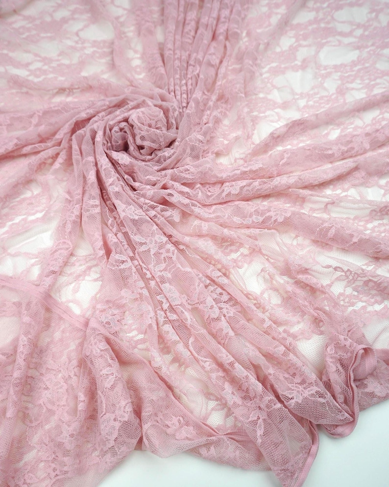 May include: A close-up shot of a delicate, light pink lace fabric. The fabric is draped and swirled, creating a soft, textured appearance. The lace features a floral pattern, adding to its romantic and feminine aesthetic. Ideal for bridal wear or craft projects.