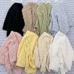 Op de afbeelding: Een verzameling stoffen met textuur in verschillende kleuren, waaronder zwart, taupe, salie, blauw, stoffig roze, geel, cr&egrave;me en ivoor. De afbeelding toont de stof met de kleurnamen boven en onder elk stuk.
