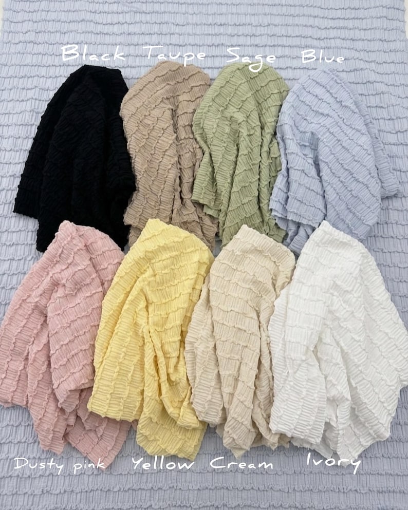 Op de afbeelding: Een verzameling stoffen met textuur in verschillende kleuren, waaronder zwart, taupe, salie, blauw, stoffig roze, geel, cr&egrave;me en ivoor. De afbeelding toont de stof met de kleurnamen boven en onder elk stuk.